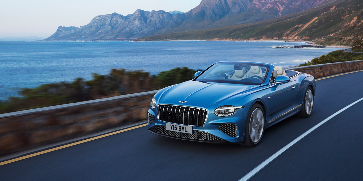 Emil Frey Exclusive Cars GmbH | Bentley München New Bentley Continental GTC Azure convertible in Light Windsor blue paint with 22 inch Azure wheels and chrome grill, driving dynamically by the sea