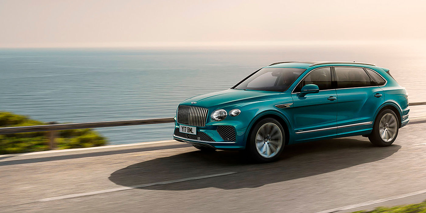 Emil Frey Exclusive Cars GmbH | Bentley München Front side angled view of a Topaz blue Bentley Bentayga EWB Azure, cruising alongside the sea.