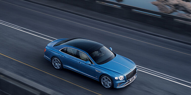 Emil Frey Exclusive Cars GmbH | Bentley München New Bentley Flying Spur Azure sedan in Light windsor blue paint by Mulliner, with 22 inch Azure wheels seen from overhead driving dynamically on a country road