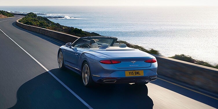 Emil Frey Exclusive Cars GmbH | Bentley München New Bentley Continental GTC Azure convertible rear view in Light Windsor blue paint with 22 inch ten spoke wheels driving dynamically on an ocean road in the sunshine