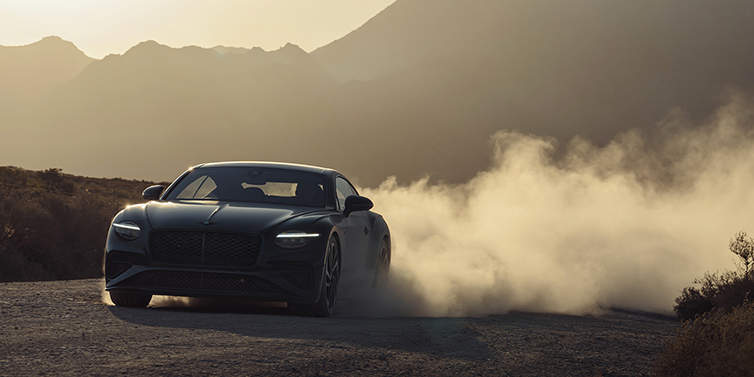 Emil Frey Exclusive Cars GmbH | Bentley München Bentley Continental GT coupe in Thunder paint front 3/4 driving with dust trail at sunset